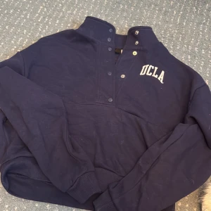 Mörkblå UCLA sweatshirt från H&M Divided - Säljer en mörkblå sweatshirt från H&M Divided i samarbete med UCLA. Tröjan har tryckknappar vid halsen, stor vit UCLA-logga på ryggen och mindre tryck fram. Ribbad mudd vid ärmslut och nederkant. Perfekt för dig som gillar college-vibe!