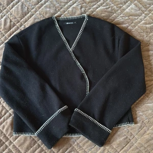 Svart omlottkofta från Gina Tricot - Svart omlottkofta från Gina Tricot med vit dekorativ söm längs kanterna. Koftan har långärmade ärmar och en stilren, enkel design som passar perfekt till jeans eller kjol. Materialet känns mjukt och har en lite tjockare känsla, perfekt för kyliga dagar.