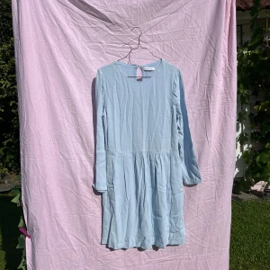 Ljusblå klänning från Samsøe Samsøe - Supersöt ljusblå klänning från Samsøe Samsøe i mjuk viskos. Modellen är kort med långärmade ärmar och har en rund halsringning med snygg öppning bak. Kjoldelen är lätt rynkad för en chill och flowy vibe. Perfekt för dig som gillar enkel och stilren design.