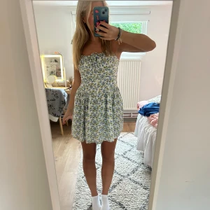 Blommig kort klänning - Supersöt sommarklänning med blommigt mönster i blått, grönt och gult på vit botten. Klänningen är kort, har smala axelband med knyt och en rynkad överdel med liten rosett framtill. Materialet känns lätt och luftigt, köpt i london