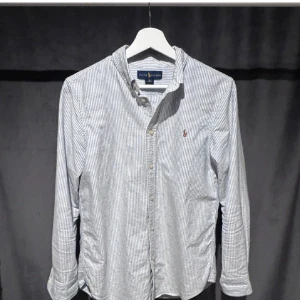 Randig skjorta från Ralph Lauren - Klassisk skjorta från Ralph Lauren i vitt och blått med smala ränder. Skjortan har lång ärm, knappar framtill och den ikoniska broderade loggan på bröstet. Tillverkad i mjuk bomull och passar perfekt till en stilren look.