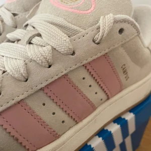 Adidas Campus i beige och rosa mocka - Snygga Adidas Campus sneakers i beige mocka med klassiska rosa ränder på sidorna. Skorna har vit sula, breda vita skosnören och broderad logga i guld och rosa på plösen. Perfekta för dig som gillar en clean och cool look med en touch av färg. Endast använd ett fåtal gånger (ca 3)
