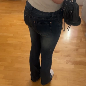 Mörkblå bootcut jeans med knappar - Snygga mörkblå jeans med bootcut passform och tre dekorativa knappar i midjan. Jeansen har klassiska fickor bak och fram, samt lätt slitning på låren för en cool look. Perfekta för dig som gillar en tidlös men trendig stil.