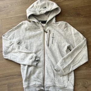 Grå hoodie från Sail Racing med dragkedja - Ljusgrå hoodie från Sail Racing med hel dragkedja och extra bröstficka med dragkedja. Broderad logga på bröstet och huva med snygg passform. Perfekt för chill dagar eller när du vill ha en sportig look.