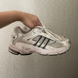 Adidas vita sneakers med mesh och snörning - Säljer ett par klassiska Adidas sneakers i vitt med silver- och grå detaljer. Skorna har ventilerande meshpaneler, tre ikoniska ränder på sidorna och platt sula. Perfekta för dig som gillar retrostil och sportig vibe. Skorna har rund tå och snörning framtill. Har använts några gånger men är i ganska bra skick, ingen lukt och det gårsäkert att tvätta bort de lite smutsiga!