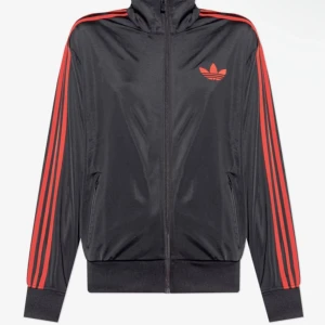 Svart Adidas vindjacka med röda ränder - Snygg svart vindjacka från Adidas med klassiska röda ränder längs ärmarna och röd logga på bröstet. Jackan har dragkedja framtill, hög krage och ribbade muddar. Perfekt för dig som gillar sportig stil och streetwear. Köpt för 829 vet inte storlek 