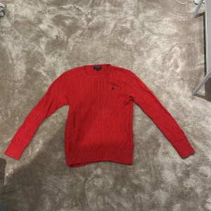 Röd kabelstickad tröja Polo Ralph Lauren - Skick: 9/10, knappt använd. Säljer en röd kabelstickad tröja från Polo Ralph Lauren med klassisk rund hals och broderad marinblå logga på bröstet. Tröjan har långa ärmar och ribbade muddar vid ärmslut och nederkant. Perfekt för dig som gillar preppy stil och vill ha något snyggt och bekvämt.