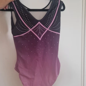 Lila och svart gymnastikdräkt - Snygg gymnastik dräkt i lila och svart med coola geometriska linjer i rosa och glittrande detaljer framtill. Materialet är mjukt och stretchigt syntet, perfekt för träning Modellen har en sportig och modern look med breda axelband och v-ringad front. Säljer då den inte används längre