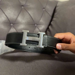 Svart skärp med H-spänne - Snyggt svart skärp med ikoniskt H-formad metallspänne i silver. Bältet är gjort i äkta skinn med tydliga vita sömmar längs kanterna. Perfekt accessoar för att lyfta din outfit med en lyxig touch.