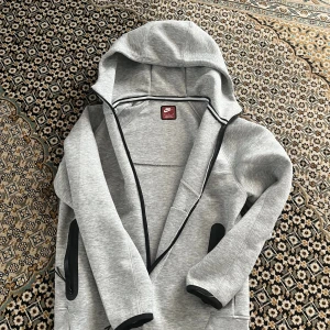 Grå Nike hoodie med dragkedja M - Snygg grå hoodie från Nike i storlek M. Tröjan har hel dragkedja framtill, svarta detaljer vid fickor och muddar samt en klassisk huva. Perfekt för chill eller träning. Materialet är mjukt och känns sportigt. (Priset kam diskuteras)