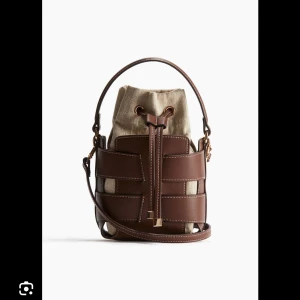 Hm bucketväska  - BUDA VID INTRESSE!Snygg bucket bag i brun skinn med kontrasterande beige bomullstyg. Väskan har både handtag och axelrem samt cool snörning upptill med gulddetaljer. 
