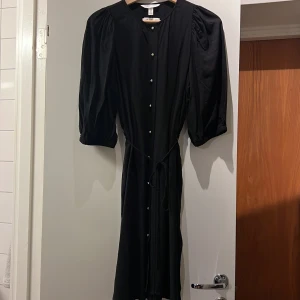 Svart långklänning från H&M - Svart långklänning från H&M i 100% LivaEco viskos. Klänningen har puffiga trekvartsärmar, knappar framtill och ett knytband i midjan för snygg passform. Enkel och stilren design som passar perfekt när du vill känna dig extra chic.