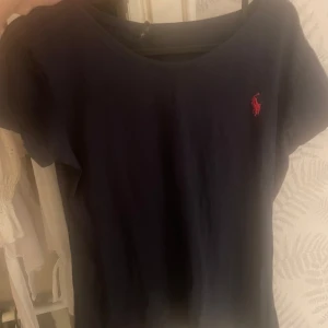 Mörkblå t-shirt från Ralph Lauren - Snygg mörkblå t-shirt från Ralph Lauren med klassisk röd logga broderad på bröstet. T-shirten har rund halsringning och korta ärmar, perfekt för en clean och stilren look.