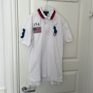Vit Polo Ralph Lauren t-shirt USA - Vit t-shirt från Polo Ralph Lauren med broderad USA-flagga, stor marinblå polospelare på bröstet och siffran 3 på ärmen. Kragen har röda och marinblå detaljer. Klassisk och sportig stil, perfekt för dig som gillar preppy vibes.