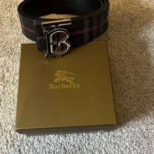 B rutigt svart bälte med B-spänne - Säljer ett snyggt svart bälte från B med klassiskt rutigt mönster i rött och grått. Bältet har ett stort metallspänne med Bs ikoniska B-logga och är gjort i exklusivt skinn. Perfekt accessoar för att lyfta din outfit.