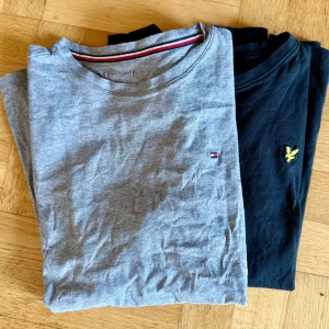 T-shirt bundle stl 170: 2 långärmade tröjor Tommy Hilfiger & Lyle & Scott - Två stilrena långärmade tröjor: en grå från Tommy Hilfiger med diskret logga på bröstet och en svart från Lyle & Scott med gul broderad logga. Båda är i mjuk bomull och har klassisk passform. Perfekta för en clean och enkel look.