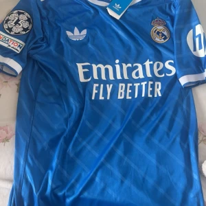 Real Madrid Bellingham blå Adidas tröja - Blå Real Madrid fotbollströja från Adidas med Bellingham och nummer 5 på ryggen. Tröjan har vita detaljer, klubbmärke, sponsortryck och UEFA-märke på ärmen. Tillverkad i lätt och ventilerande material, perfekt för match eller träning.