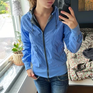 Blå Nike vindjacka med huva - Snygg blå vindjacka från Nike med huva och dragsko. Jackan har hel dragkedja framtill, ribbade muddar och svart innerfoder. Perfekt för dig som vill ha en sportig och stilren look. Passar till jeans eller träningskläder.