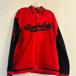 Röd och svart bomberjacka från FSBN - Snygg bomberjacka från FSBN i rött och svart med broderad text 'Reckless' på bröstet och siffran 9 på ärmen. Jackan har knappar framtill, ribbade muddar och kontrastfärgade ärmar. Perfekt för dig som vill sticka ut med en sportig och cool stil.