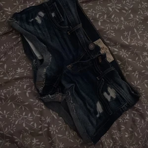 Mörkblå jeansshorts från Hollister - Snygga mörkblå jeansshorts från Hollister med låg midja och slitna detaljer. Shortsen har klassiska fem fickor och coola broderade sömmar på bakfickorna. Perfekt för dig som gillar en avslappnad och trendig stil.