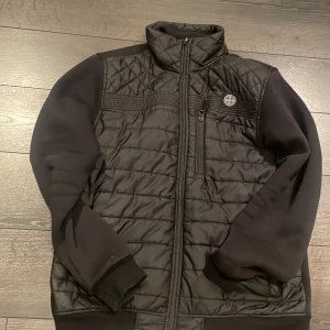 Svart quiltad jacka från Stone Island - Svart quiltad jacka från Stone Island med hög krage och dragkedja framtill. Jackan har en bröstficka med dragkedja, ribbade muddar och diskret logga på bröstet. Den är fin och brakvalite och är fräsch utan några fläckar och hål 
