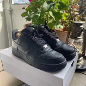 Svarta Nike Air Force 1 sneakers - Säljer ett par klassiska helsvarta Nike Air Force 1 sneakers i skinn. Skorna har låg profil, rund tå, perforerad tåbox och svart sula. De har snörning och är perfekta för dig som gillar en clean och tidlös look.