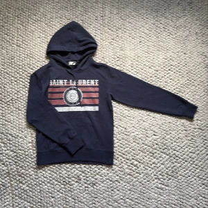 Saint Laurent hoodie - Marinblå hoodie från Saint Laurent med stort vintageinspirerat tryck i rött och vitt framtill. Klassisk huva med snörning och känguruficka. Långärmad och i mjuk bomull, perfekt för en chill och snygg look. Tags tillkommer och fraktar samma dag vid köp! Tveka inte på frågor!