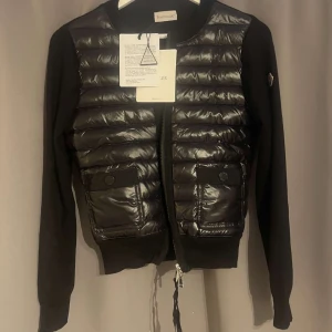 Svart quiltad cardigan jacka Moncler (Helt ny) - Snygg svart cardigan jacka från Moncler med quiltad, glansig framsida och två fickor med knappar. Långärmad med dragkedja framtill och Moncler-logga på ärmen. Perfekt för dig som vill ha en stilren och trendig look.