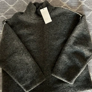 Mörkgrå polotröja från ZARA - Mörkgrå polotröja från ZARA med oversized passform och lång ärm. Tröjan har en hög krage och är tillverkad i ett mjukt, stickat material som känns extra mysigt. Diskret vit detalj längs axlarna ger en cool touch. Perfekt för kalla dagar.