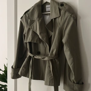 Beige trenchcoat från Bershka XS - Kort khaki trenchcoat från Bershka i storlek XS. Jackan har bred krage, knytskärp i midjan, knappar framtill och axelklaffar. Perfekt för lager-på-lager och snygg till både jeans och kjol. Materialet känns lätt och passar bra till vår och höst. Aldrig använt!