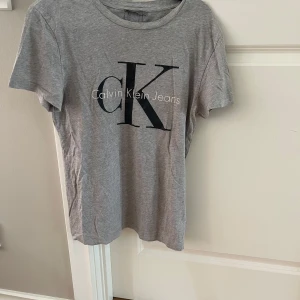 Grå Calvin Klein Jeans t-shirt S - Snygg grå t-shirt från Calvin Klein Jeans i storlek S. Klassisk rund hals och korta ärmar. Stor CK-logga i svart och vitt framtill ger en clean och modern vibe. Mjuk bomull som är skön mot huden och passar perfekt till jeans eller shorts.