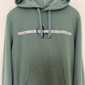 hoodie från Calvin Klein  - Snygg grön hoodie från Calvin Klein i storlek M. Tröjan har huva med snörning, magficka och tryckt logga samt Calvin Klein-band över bröstet. Perfekt för dig som gillar stilrena och bekväma plagg.