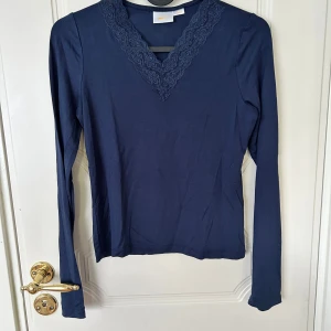 Marinblå långärmad topp Gina Tricot - Mörkblå långärmad topp från Gina Tricot med v-ringning och snygg spetsdetalj runt halsen. Tillverkad i mjuk bomull, perfekt för dig som gillar en enkel men ändå söt stil. Passar till både jeans och kjol.