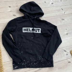 Svart Helmut Lang hoodie med tryck - Svart hoodie från Helmut Lang med stor vit logga på bröstet. Tröjan har huva med snörning, känguruficka och coola vita färgstänk nertill. Perfekt för dig som gillar streetstyle och vill sticka ut lite extra. Den är i fint skick.