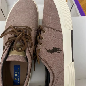 Bruna sneakers från Polo Ralph Lauren - Snygga bruna sneakers från Polo Ralph Lauren med klassisk logga på sidan. Skorna har snörning, vit platt sula och är tillverkade i bomullstyg med bruna mockaliknande skosnören. Perfekta för en avslappnad och stilren look nu i höst! Pris kan diskuteras!💕