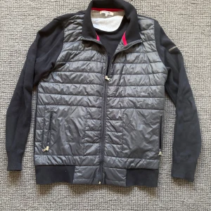 Svart quiltad jacka från Iceland L - Säljer en svart quiltad jacka från Iceland i storlek L. Jackan har stickade ärmar och rygg, quiltad framsida i syntetmaterial och dragkedja framtill. Två fickor med dragkedja och hög krage med röd detalj på insidan. Perfekt för kyliga dagar.