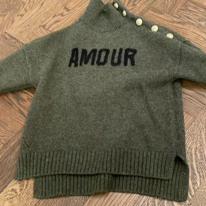 Olivgrön stickad tröja Zadig & Voltaire - Olivgrön stickad tröja från Zadig & Voltaire med svart 'AMOUR'-text framtill. Hög krage och snygga guldfärgade knappar längs ena axeln. Ribbstickade muddar och slits i sidorna ger en chill vibe. Perfekt för lager-på-lager under kalla dagar.strl 134-140