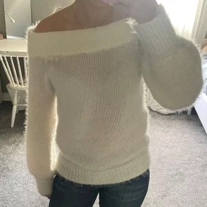 Offshoulder fluffig vit stickad tröja - Supermjuk och fluffig vit stickad tröja med offshoulder-modell. Tröjan har bred ribbad kant upptill och långa ärmar. Perfekt för dig som gillar en cozy och trendig look med lite oversized känsla.