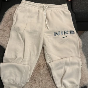 Vita/beiga mjukisbyxor från Nike stl M - Snygga vita mjukisbyxor från Nike i storlek M med broderad logga på ena benet. Byxorna har resår och snörning i midjan samt muddar vid bensluten. Tillverkade i mjukt material som är skönt mot huden. Perfekta för chill eller träning. Använt ett fåtalet gånger. 