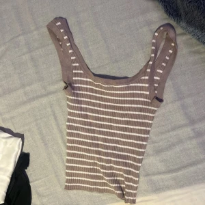 Randig ribbad linnetopp i beige och vit - Superfin ribbad linnetopp med breda axelband i beige och vitt. Toppen har ett randigt mönster och är stretchig med en figurnära passform. Perfekt för varma dagar och enkel att styla med jeans eller kjol.