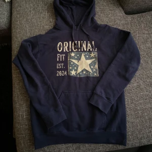 Mörkblå hoodie med stjärntryck - Snygg mörkblå hoodie från Original Fit med stort stjärntryck och text på bröstet. Tröjan har huva och en klassisk känguruficka framtill. Perfekt för en avslappnad och cool stil.