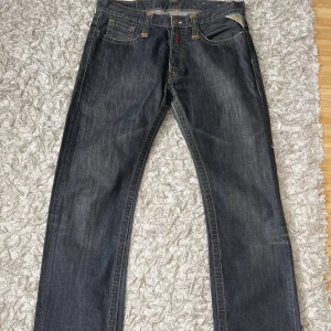 Mörkgrå jeans från Replay - Säljer ett par mörkgrå jeans från Replay med klassisk femficksdesign, raka ben och snygga kontrastsömmar. Jeansen har knäppning med knappar och Replay-logga på bakfickan samt läderpatch i midjan. Perfekta för dig som gillar en tidlös och clean look.