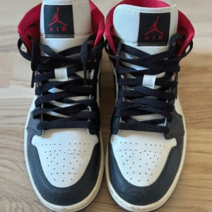Nike Air Jordan 1 Mid svart/vit/röd - Snygga Nike Air Jordan 1 Mid sneakers i svart, vitt och rött. Klassisk design med svart swoosh, perforerad tå och röd vadderad häl. Skorna har snörning och är tillverkade i skinn och syntet. Perfekta för dig som gillar streetstyle och ikoniska sneakers.