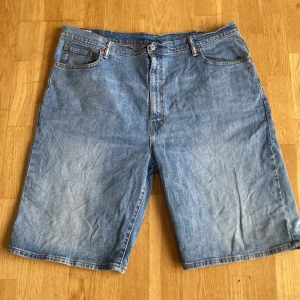 Levi's ljusblå jeansshorts - Säljer ett par klassiska ljusblå jeansshorts från Levi's med fem fickor, knapp och dragkedja framtill samt bälteshällor. Shortsens raka passform och slitna denim ger en chill vibe, perfekt för varma dagar. Levi's ikoniska läderpatch bak på midjan.