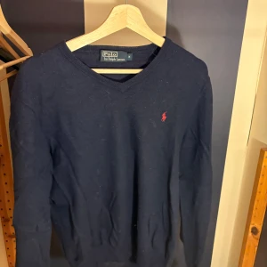 Mörkblå stickad tröja Polo Ralph Lauren - Snygg mörkblå stickad tröja från Polo Ralph Lauren med klassisk v-ringning och rött broderat logomärke på bröstet. Tröjan har långa ärmar och ribbade muddar vid hals, ärmslut och nederkant. Perfekt för dig som gillar stilren och enkel design.