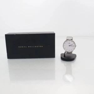 Silverfärgad klocka från Daniel Wellington - Stilren klocka från Daniel Wellington med rund vit urtavla och silverfärgat mesharmband i metall. Minimalistisk design med tunna markeringar och DW-logga på urtavlan. Perfekt accessoar för en clean look. Behöver ett nytt batteri. 