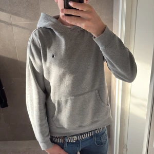 Grå hoodie från Polo Ralph Lauren  - Snygg grå hoodie från Polo Ralph Lauren . Klassisk design med huva, känguruficka och broderad logga på bröstet. Mjuk och skön bomullsmix, perfekt för chill dagar. Långärmad och med normal passform.