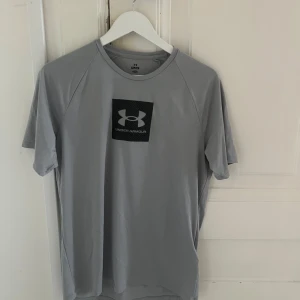 Grå t-shirt från Under Armour - Snygg grå t-shirt från Under Armour med svart logga på bröstet. T-shirten har rund hals och korta ärmar, perfekt för träning eller chill. Materialet är mjukt och lätt polyester som andas bra. Enkel design med sportig vibe.