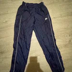 Marinblå Nike träningsbyxor XS  - Snygga marinblå träningsbyxor från Nike med vita detaljer längs benen och broderad logga på låret. Byxorna har resår i midjan och är tillverkade i ett lätt och syntetiskt material som andas. Perfekta för dig som gillar sportig stil.