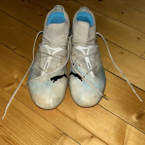 Puma vita fotbollsskor med rosa sula - Snygga fotbollsskor från Puma i vitt med svarta loggor och coola blå detaljer. Skorna har snörning, stickad ovandel och en grym rosa sula med dobbar för bästa grepp på planen. Perfekta för dig som vill sticka ut på matchen!